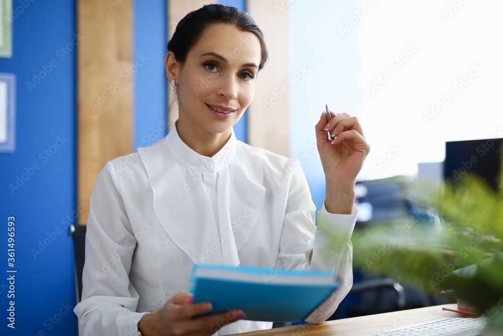 Portrait of attractive office worker sitting with blue folder and silver pen. Personal cabinet with blue wall. Charming lady in stylish suit. Successful career and business concept Portrait of attractive office worker sitting with blue folder and silver pen. Personal cabinet with blue wall. Charming lady in stylish suit. Successful career and business concept