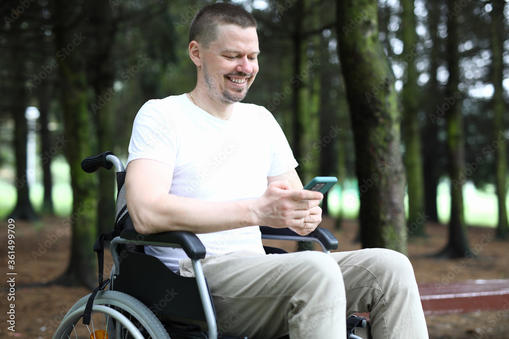 Obraz premium Portrait of cheerful happy person sitting in wheelchair and using smartphone for fun. Laughing man in white shirt having fun in forest. Smiling adult enjoying life on nature. Disabled people concept