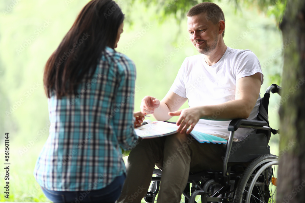 Portrait of smiling man sitting in wheelchair with papers. Handsome middle aged person in white shirt. Woman holding folder with documents. Working moments. Business and disabled people concept Portrait of smiling man sitting in wheelchair with papers. Handsome middle aged person in white shirt. Woman holding folder with documents. Working moments. Business and disabled people concept