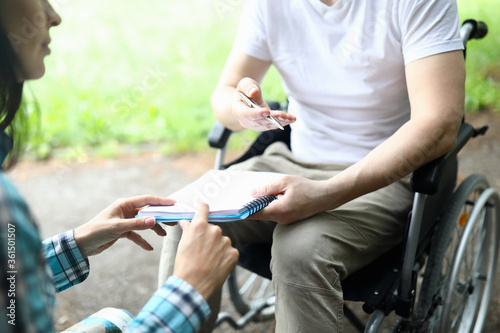 Close-up of man sitting in wheelchair and holding paper for signing. Woman showing place for signature. Business moment and meeting in park. Disabled people concept