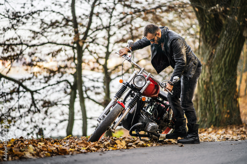 Bearded brutal man in sunglasses and leather jacket sitting on a motorcycle on the road in the forest Bearded brutal man in sunglasses and leather jacket sitting on a motorcycle on the road in the forest