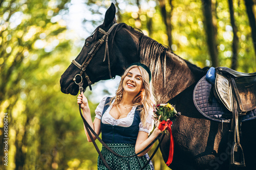 Pretty blonde in traditional dress  walking with big black horse in the forest