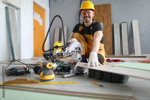 Portrait of cheerful smiling worker cutting wooden laminate. Qualified male wearing yellow helmet and white protective gloves. Happy craftsman and drill machine. Renovation concept