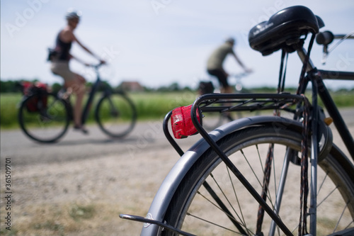 Two blurry cyclists cycle past a parked bicycle on a country road on a summer day. Focus on mudguard with red rear light