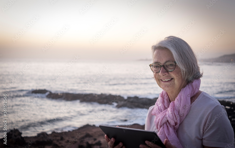 Cute and smiling senior woman gray hair sitting near the beach surfing the net with tablet. Dusk light and horizon over water. Happy retirement concept