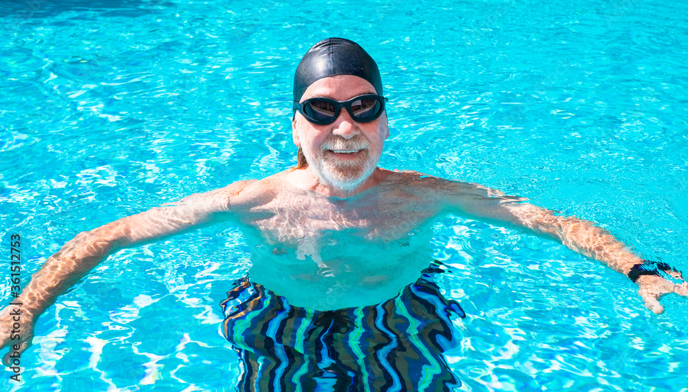Senior bearded man smiling in the sun swims in the outdoor pool, wearing goggles and swimming cap, active and happy people in retirement concept