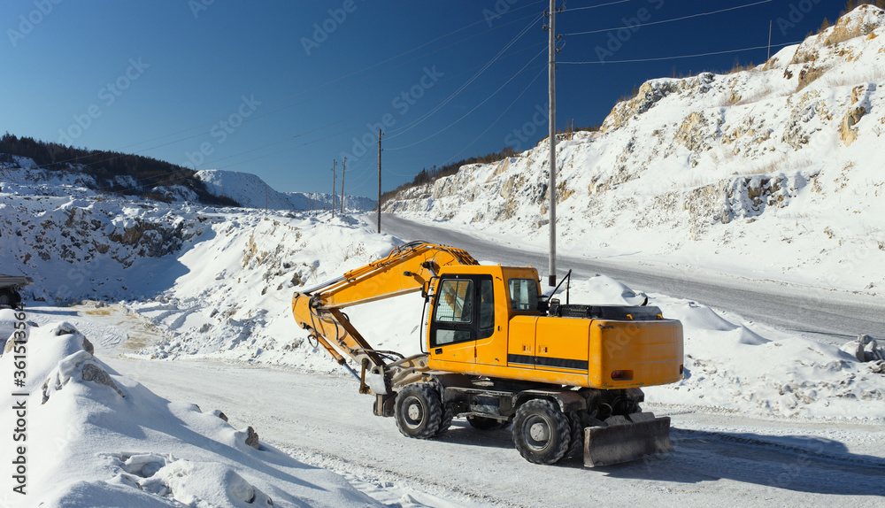 Excavator with hydraulic hammer in a limestone quarry on the background of snow-covered rocks and blue sky, panorama. Excavator with hydraulic hammer in a limestone quarry on the background of snow-covered rocks and blue sky, panorama.