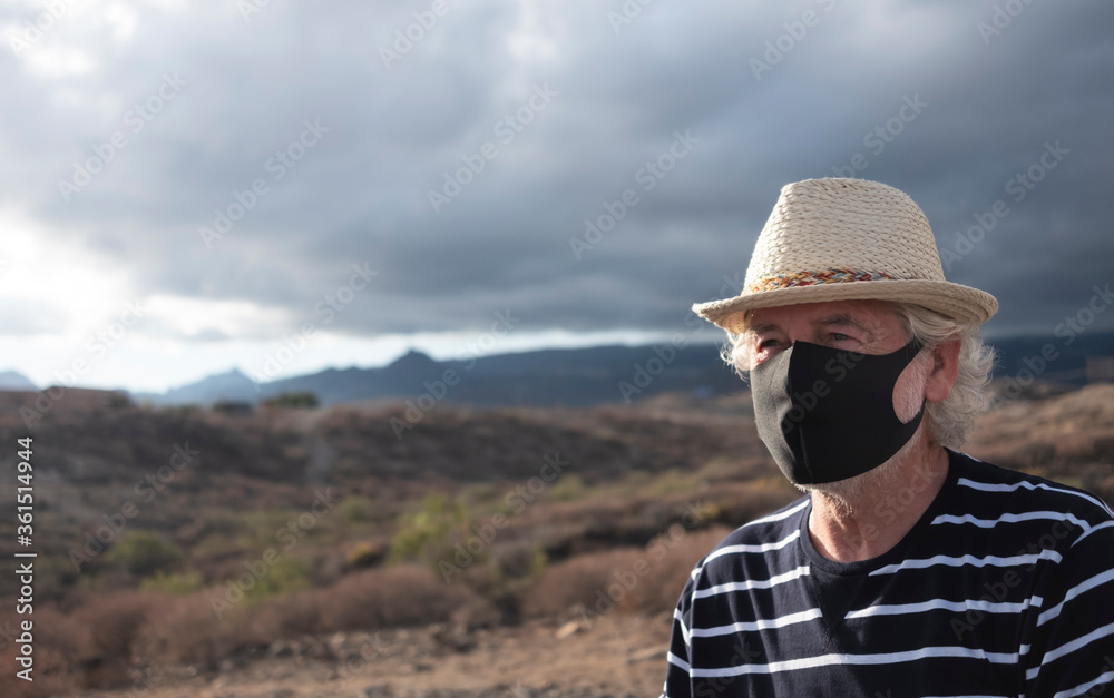 Serious senior man with straw hat and blue striped shirt in arid landscape wearing a mask because of the Covid-19 coronavirus. Mountains and overcast sky behind him - active retirement concept