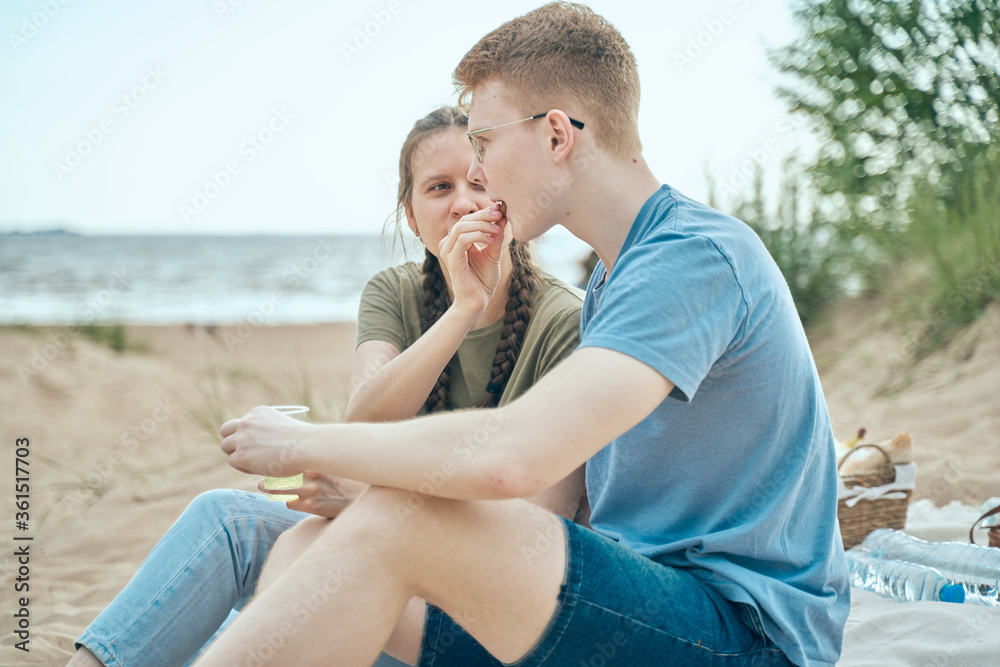 Picnic on beach with food and drinks. Young girl feeds boy, sitting on sand