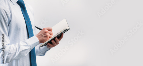 The guy writes in a black notebook. A man on a white background with a blue tie in a white shirt. Note in a notebook. Panoramic photo with an object from the left and a place for an inscription..