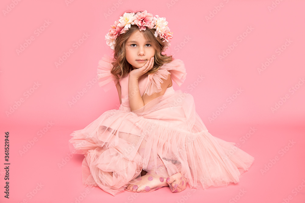 Studio photo of a small beautiful girl with a wreath of flowers on her head and curls of hair. Background, accessories, pink dress. Hairdressing salon. Children's cosmetic.