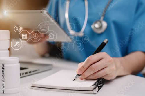 Male medicine doctor, physician or practitioner in lab room writing on blank notebook and work on laptop computer with medical stethoscope on the desk.