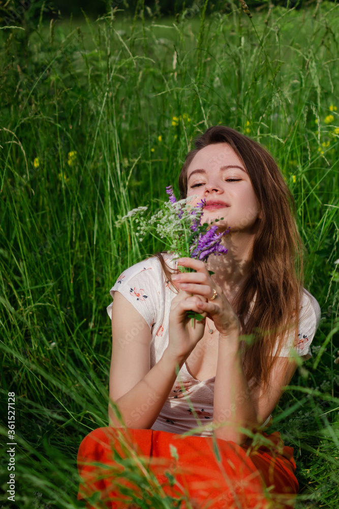 Young brunette girl in white top and red skirt sitting in green field grass in countryside smelling small bouquet of field flowers in hands with closed eyes and enjoy nature and rural life. Young brunette girl in white top and red skirt sitting in green field grass in countryside smelling small bouquet of field flowers in hands with closed eyes and enjoy nature and rural life.