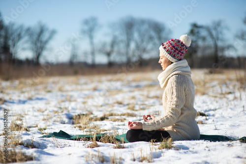 Young athletic woman sitting and meditatingin the yoga pose on the snowy field during winter or early spring walk