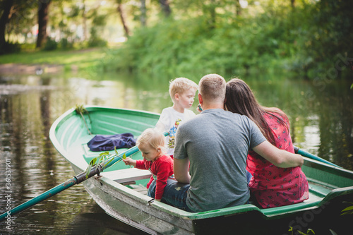The family is floating on the lake in a boat with oars in the park, dad hugs mom, the girl looks into the water and holds a twig. Back view.