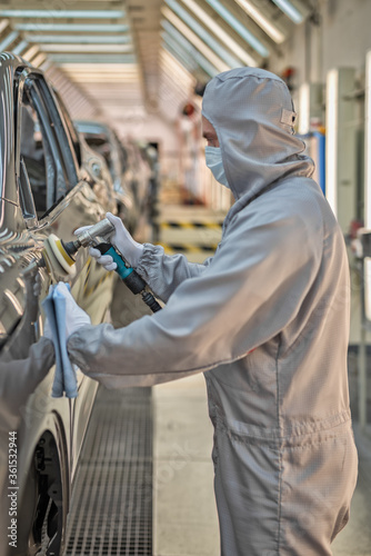 An employee of the car body paint shop with a medical mask on his face polishes the painted surface