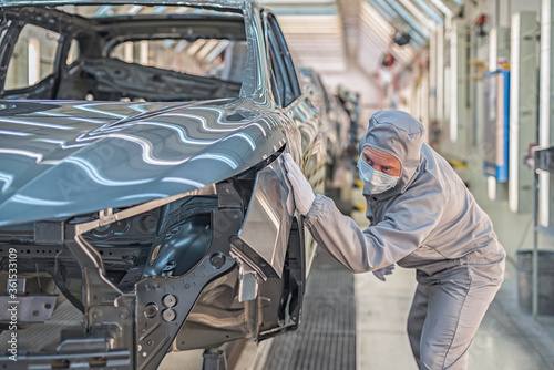 An employee of the car body paint shop with a medical mask on his face checks the quality of the painted surface