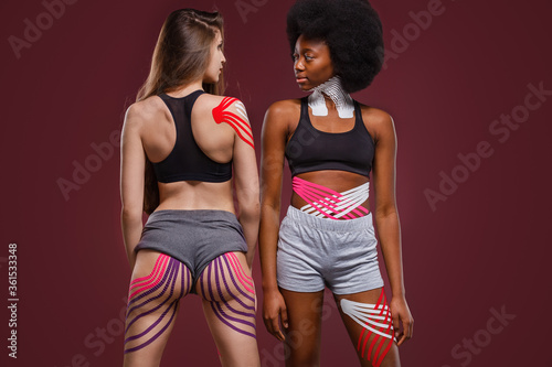 Kinesiology taping. Two young female athletes on red background with kinesiology tape on neck,shoulder, khee, tummy and hips. Fat lose, cellulite removal, sport physical therapy,recovery concept. photo