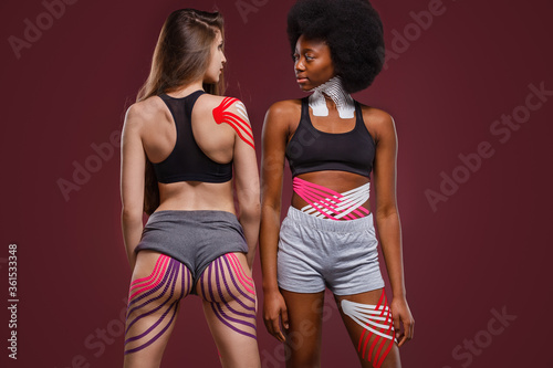 Kinesiology taping. Two young female athletes on red background with kinesiology tape on neck,shoulder, khee, tummy and hips. Fat lose, cellulite removal, sport physical therapy,recovery concept.