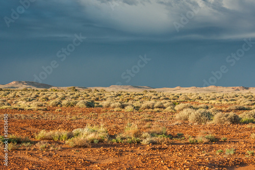 Outback landscape in South Australia with flat red ground with gravel and rocks covered with shrubs and a ridge at the horizon  against dark blue and grey sky with clouds