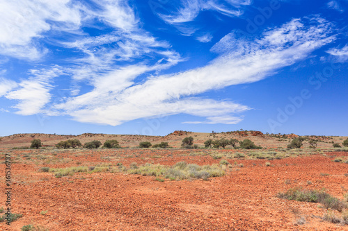Outback landscape in South Australia with flat ground with gravel and rocks, a Creek with banks covered with Gum Trees and a ridge against blue sky with light cloud cover