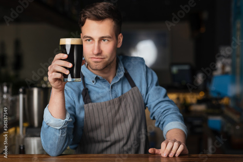 Brewer and bartender. Handsome stylish man in apron looks at glass of beer