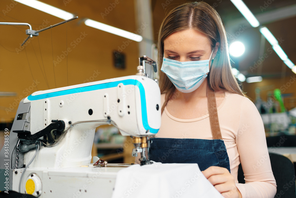 Young caucasian woman tailor working in sewing factory wearing protective medical mask