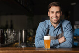 Beer traditions in modern pub. Smiling handsome guy in denim shirt leans on bar counter with glass