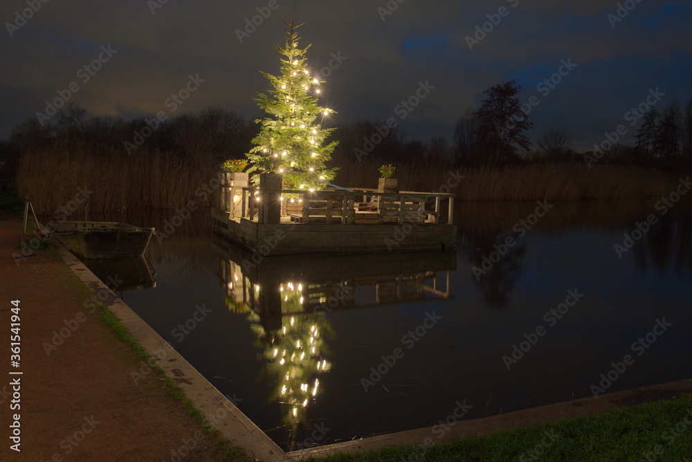 Ein beleuchteter Tannenbaum liegt auf einer kleiner Insel, gebaut aus  Holzbretter auf einem Teich. Es ist Nacht und dunkel. Hinter dem Tannenbaum  sieht man Wasserpflanzen, daneben ein kleiner Boot. Stock-Foto | Adobe