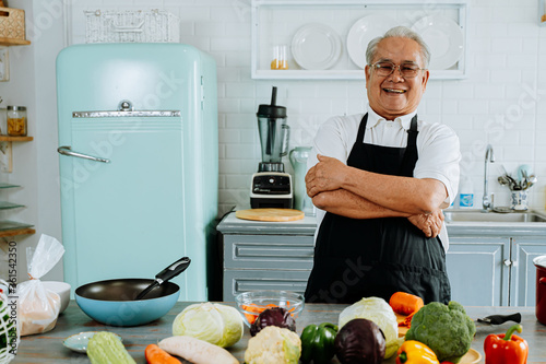 Happy senior Asian male in apron laughing and smiling for camera and having arms folded while preparing lunch in kitchen at home in arms crossed pose.