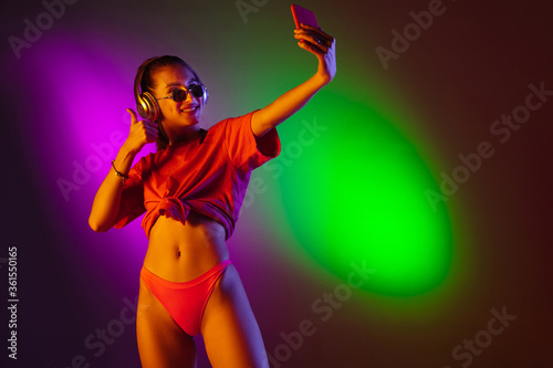 Seductive young girl' portrait isolated on bicolored neon studio background in neon. Fit sportive woman in bodysuit taking selfie. Facial expression, summer, weekend, beauty, resort concept. Vacations