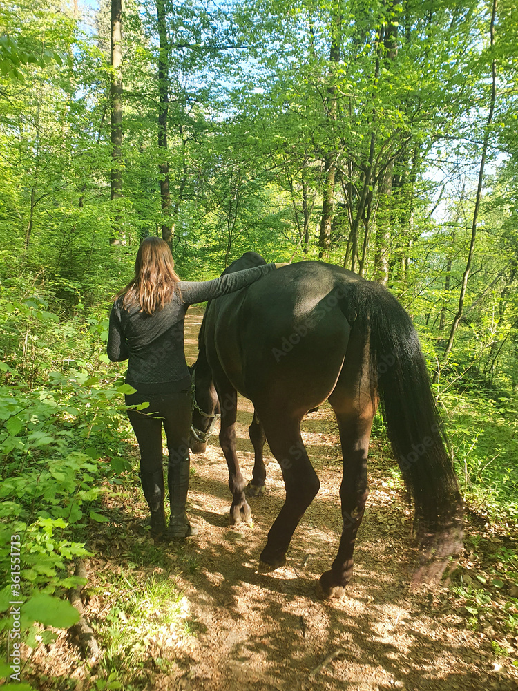 VERTICAL: Unrecognizable young woman walks down the forest trail with her horse.