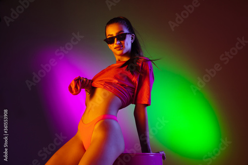 Seductive young girl' portrait isolated on bicolored neon studio background in neon. Fit sportive woman in bodysuit posing on pink barrel. Facial expression, summer, weekend, beauty, resort concept.