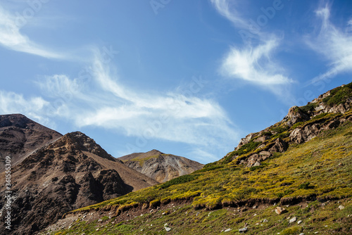 Scenic mountain landscape with beautiful cirrus clouds in clear blue sky over rocks in sunlight. Colorful highland scenery with spindrift clouds in blue clear sky above green brown rocky mountains.