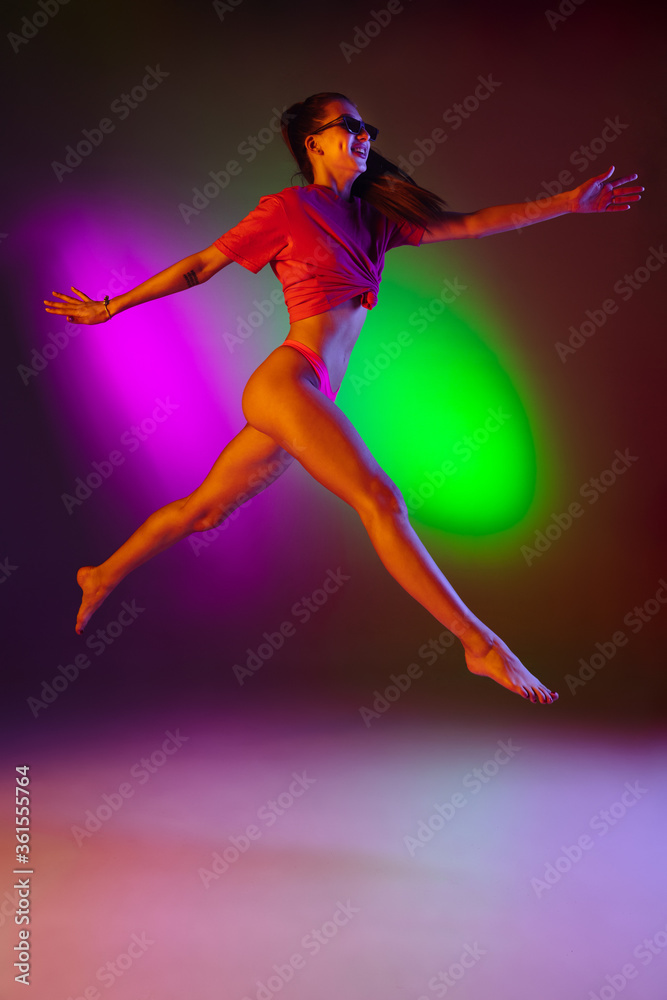 Seductive young girl' portrait on bicolored neon studio background in neon. Fit sportive woman in bodysuit running, jumping high. Facial expression, summer, weekend, beauty, resort concept. Vacations.