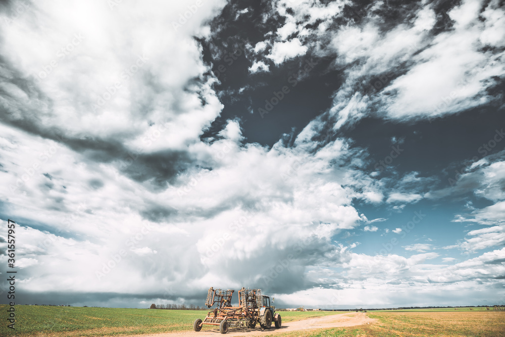 Fototapeta premium Tractor With Attached Harrow In Sunny Spring Day. Countryside Rural Road Through Field Meadow Landscape Under Scenic Dramatic Sky With Clouds Before Rain