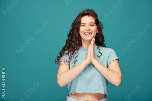 Cheerful girl with curly hair touching hands together in excitement against blue background