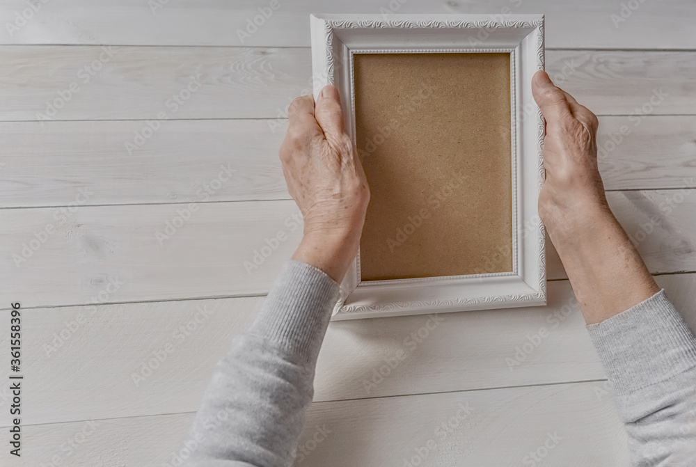 Senior wrinkled hands holding white photo frame. Elderly woman looking at the picture, light wodden background. Empty template, mock-up. Happy memories, sadness, nostalgia concept. Senior wrinkled hands holding white photo frame. Elderly woman looking at the picture, light wodden background. Empty template, mock-up. Happy memories, sadness, nostalgia concept.