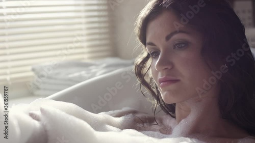 Close-up of attractive woman relaxing in bubble bath. Video. Sexy woman takes bath with foam in cozy environment on background of window