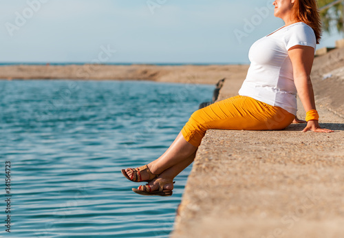 Fototapeta Naklejka Na Ścianę i Meble -  Close up portrait of a smiling overweight woman looks into the distance, sitting on a sea pier. opy space. Side view
