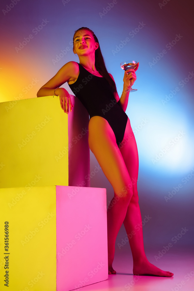 Seductive young girl on bicolored neon studio background in neon. Fit sportive woman in bodysuit fashionable posing on boxes. Facial expression, summer, weekend, beauty, resort concept. Vacations.