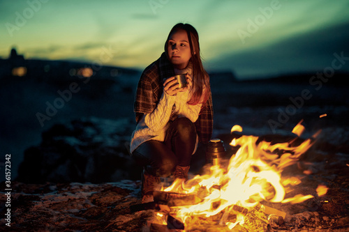 Fototapeta Naklejka Na Ścianę i Meble -  Girl-traveler warms herself in the evening at a fire in a mountain camp. A woman is sitting by the fire with hot tea in her hands.