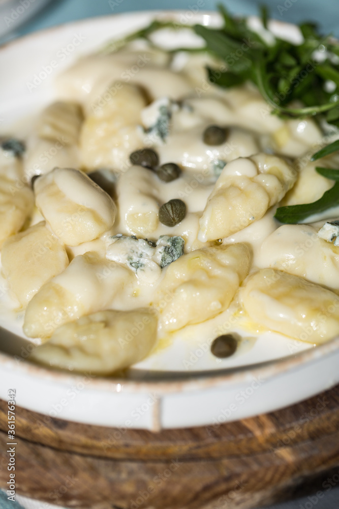 Homemade potato gnocchi with creamy gorgonzola sauce decorated with capers and arugula in white plate on wooden board.