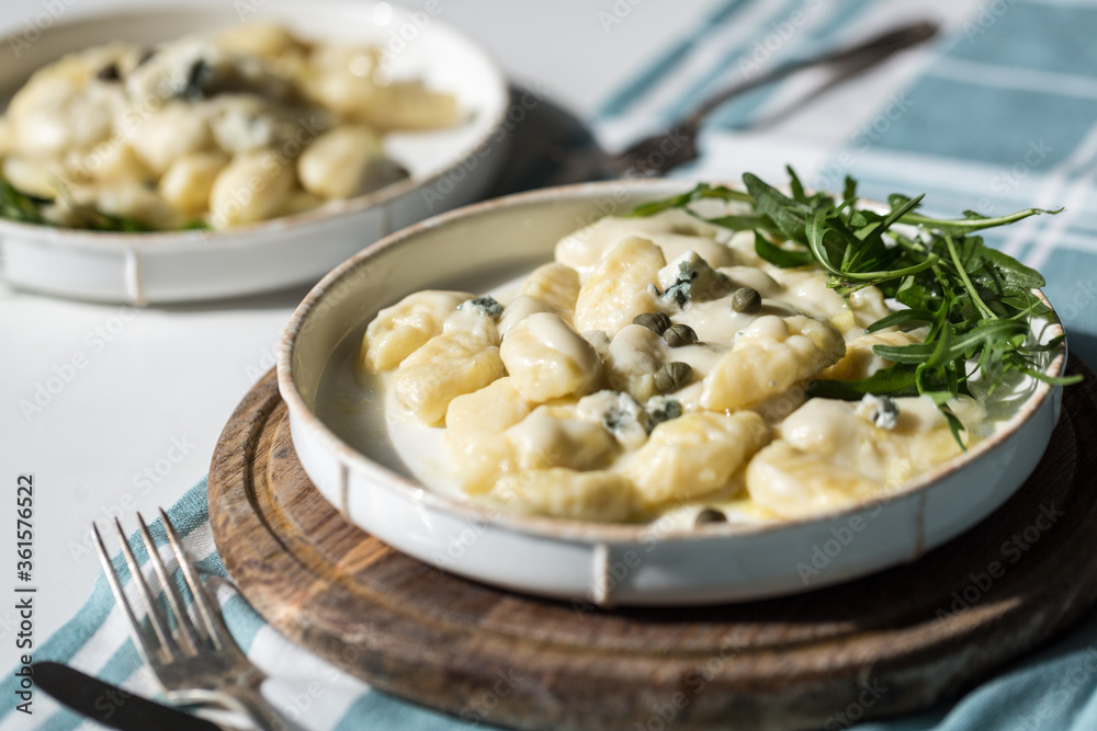 Homemade potato gnocchi with creamy gorgonzola sauce decorated with capers and arugula in white plate on wooden board.