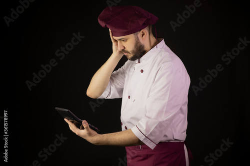 Shocked chef man in uniform holds calculator and looks on it with surprise. Сook in apron on black background. Restaurant bankruptcy, food industry crisis, business problem, liquidation, incur loss