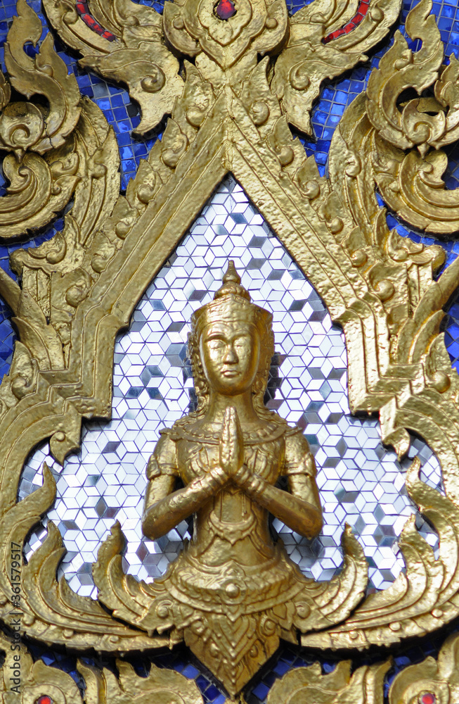 Gros plan sur un petit buste de Bouddha doré en position de prière, les mains jointes, sur fond de mosaïque en miroir, et dorures représentant des flammes ou de la végétation sur un mur peint en bleu.