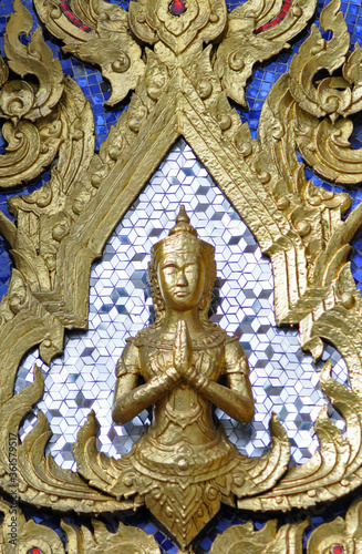 Gros plan sur un petit buste de Bouddha doré en position de prière, les mains jointes, sur fond de mosaïque en miroir, et dorures représentant des flammes ou de la végétation sur un mur peint en bleu. © Delphine