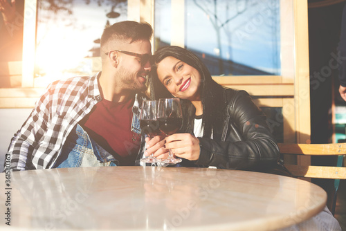 Young handsome man and his pretty stylish girlfriend are sitting at the table in the cozy cafe and holding the wine glass. Female smiling at the camera, boyfriend is looking at her. Couple looks happy