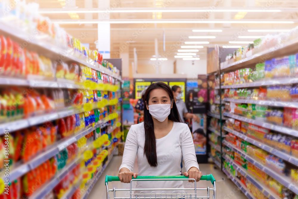 Asian woman wear face mask push shopping cart in supermarket department store. Girl looking grocery to buy  something. During coronavirus crisis or covid19 outbreak. Women wearing protective face mask