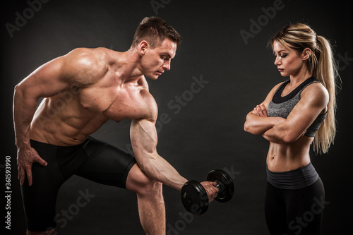 Fitness in gym  sport and healthy lifestyle concept. Couple of athletic man and woman showing their trained bodies on black background. Two bodybuilder models standing and demonstrating tight muscles.